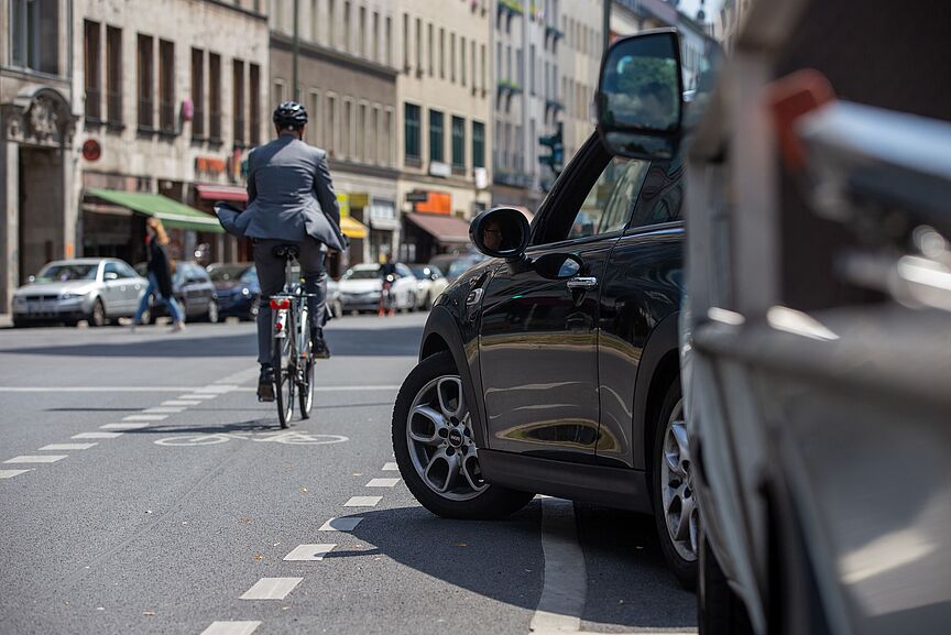 Radverkehr im Stadtverkehr Radfahrer in dunkler Kleidung mit Helm fährt auf einer Stadtstraße, im Vordergrund steht ein schwarzes Auto am Straßenrand, weitere Fahrzeuge und Gebäude im Hintergrund.