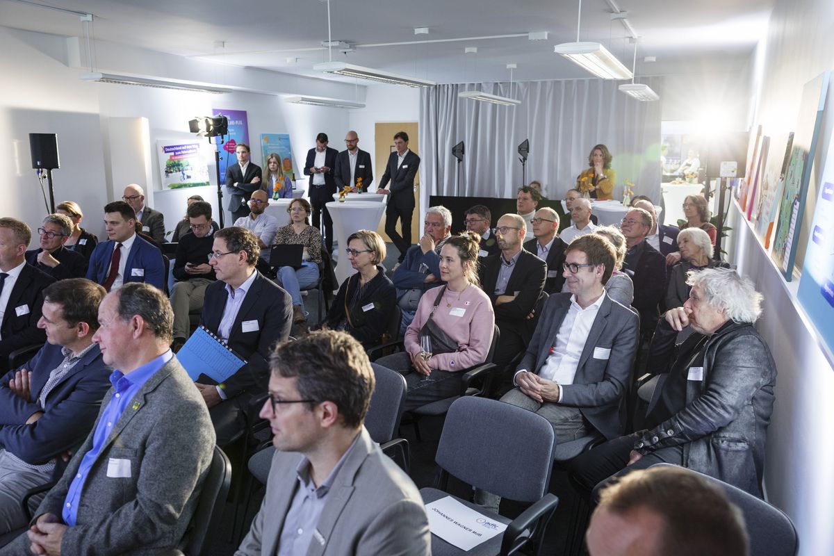 Zahlreiche Gäste sitzen auf Stühlen in einem Raum und sehen Richtung Bühne, die nicht im Bild ist. 